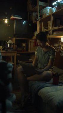 Genç Koreli bir kadının gündelik kıyafetleriyle dar bir dairede oturmuş, çizim defterinde kalemle bir şeyler çizerken ve elektrikli ocakta yemek pişirirken tam dikey görüntüleri.