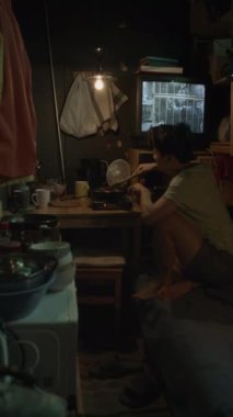 Asyalı bir kadının eski püskü bir dairede ranzada otururken, elektrikli ocakta yemek pişirirken, kürek çekerken ve akşam evde dinlenirken TV 'de film izlerken tam dikey görüntüsü.