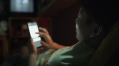 Çinli erkek ya da kadının minicik bir mikro dairede yatakta yatarken, akıllı telefon tutarken ve internette gezinirken, TV 'de film izlerken ve işten sonra evde dinlenirken omuz üstü çekimleri.