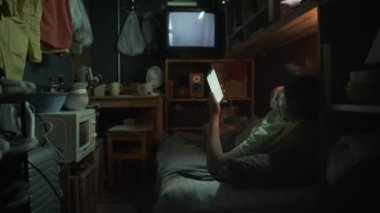 Minik, dağınık kapsül dairesindeki ranzada uyuyan, akıllı telefon kullanan, esneyen, sonra yastığa sarılan, uzanan ve TV 'de oynayan yorgun Asyalı kadın ya da erkek.