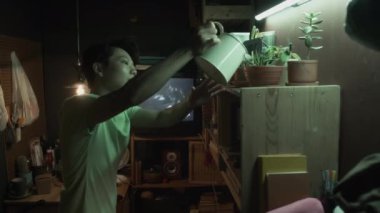 Genç, çift cinsiyetli Asyalı bir kadının, loş ışıklandırılmış mikro dairede saksıları sularken, kişisel eşyalar raflarda ve duvarlarda plastik torbalar, arka planda TV 'de film.