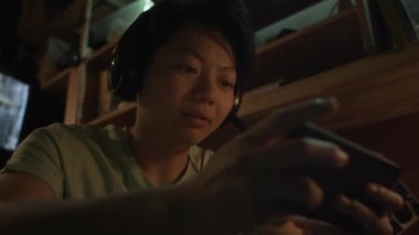 Orta ölçekli düşük açılı, kulaklıklı bir Asyalı erkek ya da kadın yatakta oturmuş akıllı telefondan video oyunu oynuyor ve sabırsızlık ve heyecanla konuşuyor.
