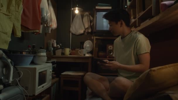 中完整镜头 雄性不育的中国女人坐在狭小的小公寓的双层床上 用智能手机 然后从微波炉中取出食物盒 放回到床上取暖 — 图库视频影像
