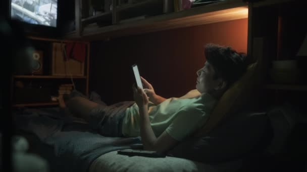 穿着T恤和短裤的中国学生躺在狭小的鞋盒公寓的双层床上 在智能手机上打字 在电视上播放恐怖片 晚上下班后放松一下 — 图库视频影像