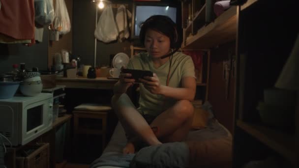 中国年轻的雌雄同体女子头戴耳机 双腿交叉地坐在凌乱的胶囊床上 迫不及待地在智能手机上玩电子游戏 下班后在家里放松 — 图库视频影像