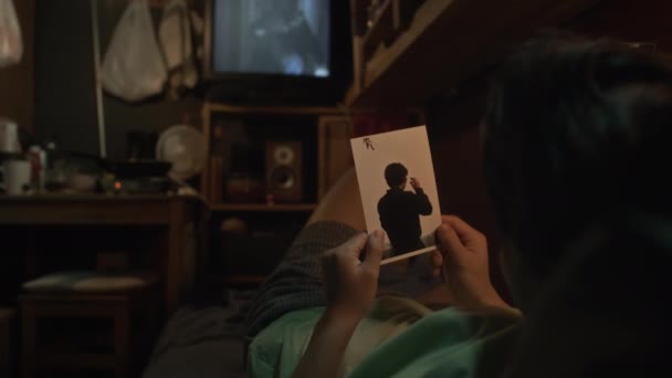 在电视前一间破旧的小公寓里 一个难以辨认的年轻亚洲人晚上躺在床铺上 拿着他男朋友的照片 看着电视 拍了照 拍了照 — 图库视频影像