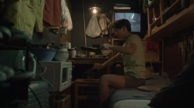 Orta boy Asyalı çift cinsiyetli bir kadının sefil bir mikro dairede ranzada oturup yemek hazırlarken, tavadan yemek yerken, arka planda TV 'de oynadığı korku filmi.