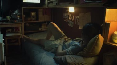 Genç Çinli ya da Koreli bir kadının gecenin geç saatlerinde karanlık bir kapsül içinde ranzada uzanmış film ya da televizyon izlerken yan yatıp uyuduğu görüntüler.