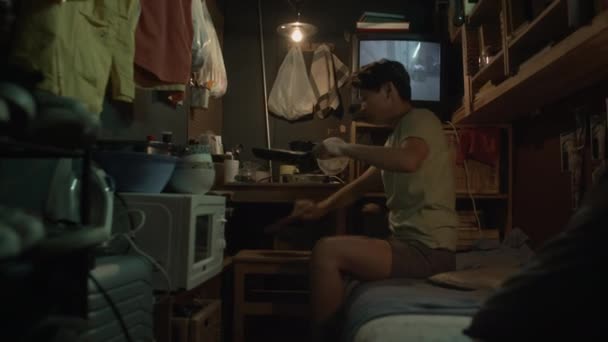 中等全景的年轻亚洲雌雄同体女子坐在破旧的小公寓里的双层床上 准备吃饭 把煎锅里的食物放进碗里 在电视上播放恐怖片 — 图库视频影像