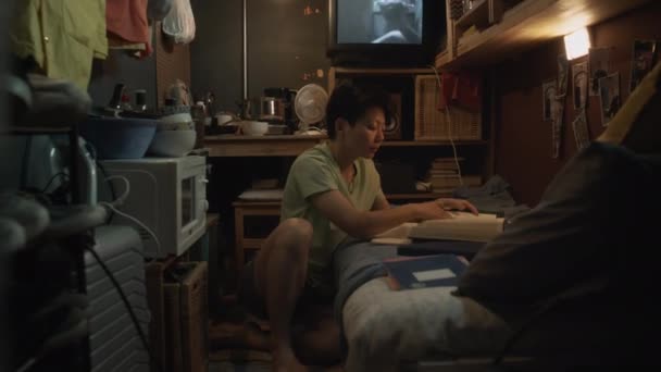 在准备高考的时候 中国年轻女子坐在凌乱的昏暗的胶囊公寓里 躺在地上 把课本放在床上 阅读和写在抄本上的全景照 — 图库视频影像