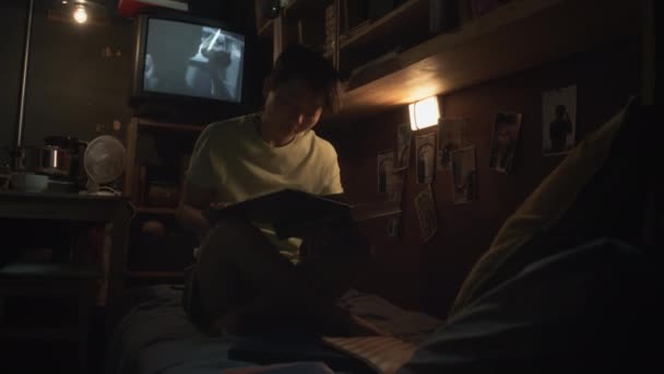 暗い小さなカプセルフラットの二段ベッドに座っている若い中国人女性の完全な映像 教科書を読み 大学の試験の準備 および背景でホラー映画を再生するテレビセット — ストック動画