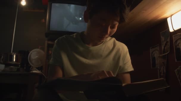 アジアの若い女性または男のミディアムクローズアップショット マイクロフラットの二段ベッドに座って 教科書を読み 大学の試験のための材料を改訂 バックグラウンドの古いテレビの映画 — ストック動画