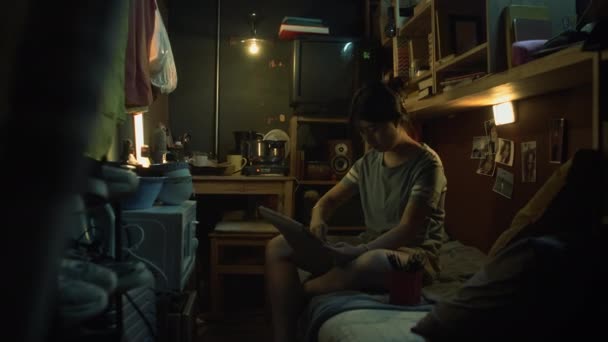 穿着T恤衫和短裤的中国小女孩坐在小公寓的双层床上 在小炉灶上做饭时 画着她那不起眼的周围环境的静物画 — 图库视频影像