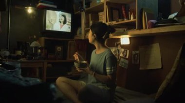 Tişörtlü genç Koreli bir kadının bacak bacak üstüne atmış bir şekilde küçük kapsül dairede oturması, yemek çubuklarıyla kızartma yemesi ve TV 'de güzellik reklamı izlemesi.