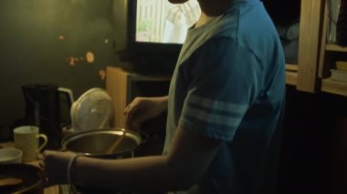 Genç Asyalı bir kadının küçük bir yatak odasında yemek pişirirken, fırında çorba pişirirken, arka planda TV 'de kredi kartı reklamında oynarken orta boy yakın çekim görüntüleri.