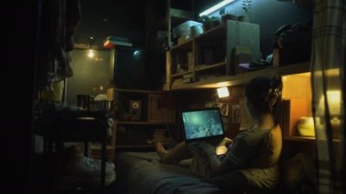 Eski püskü mikro dairede gece vakti yatakta oturan, laptop bilgisayarında video oyunu oynayan, köşe başındaki ampulün altında esrar tüttüren, tanınmamış genç bir kadının geniş arka görüntüsü.