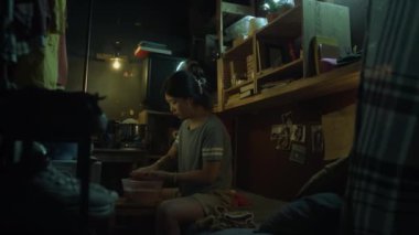 Tişörtlü ve şortlu genç Çinli bir kadın gece vakti loş ışıklı, dağınık bir mikro dairede oturuyor. Çamaşırları sabunlu su dolu plastik bir küvette elle yıkıyor.