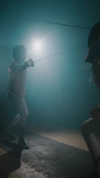 ラッパーで戦う2人のプロフェッショナルホイルフェンサーの垂直側の映像と暗い霧の部屋で優れた剣術の実演 — ストック動画