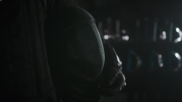暗い部屋で黒いEpeeマスクを保持する黒いユニフォームの匿名フェンサーのミスクローズアップ — ストック動画