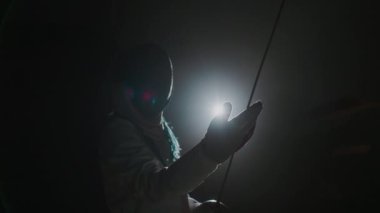 Epee maskeli folyocunun rapier 'ı tutarken ve ışığa karşı dururken bıçağına bakarken orta boy görüntüsü.