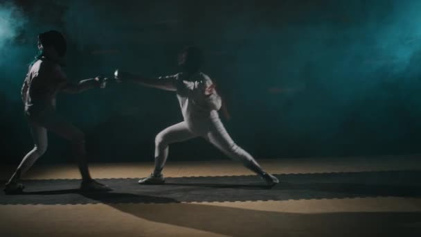 ダークスモーキーホールでホイル剣で戦う保護ギアで2人の女性間のタフなフェンシング試合またはトレーニングのサイドフルスローモ — ストック動画