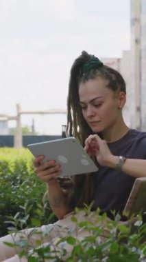 Yeşilliklerle dolu çatı katındaki bankta oturan ve tablet üzerinde çalışan bağımsız genç bir bayanın dikey görüntüleri.