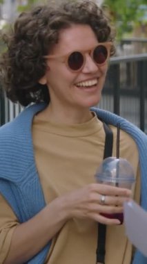 Güneş gözlüklü beyaz bir kadının arkadaşlarıyla parkta toplanıp gülüşünü gösteren dikey göğüs görüntüsü.
