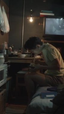 Tam dikey çekimde, Çin 'li genç bir kadın ranzada loş ışık altında oturuyor yemek çubuklarıyla yemek yiyor, akıllı telefon kullanıyor arka planda TV' de film oynuyor.