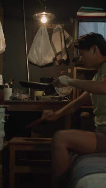年轻的中国雄雄同体者坐在简陋的小公寓的双层床上 将食物从油锅放进碗里 在电视上播放背景的电影 这些都是完整的垂直镜头 — 图库视频影像