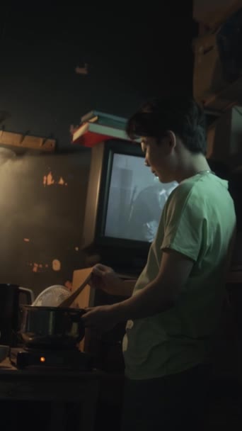 中国雄雄同体男人或女人站在灯光昏暗的胶囊公寓里 在电炉上的蒸锅里搅拌食物 在电视上看电影 晚上做饭 — 图库视频影像