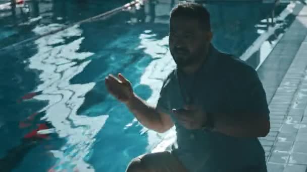 中镜头闪烁着亚洲男子教练蹲在游泳池边的镜头 向水中的运动员欢呼 做爬泳动作 看秒表 然后夸奖和展示大拇指 — 图库视频影像
