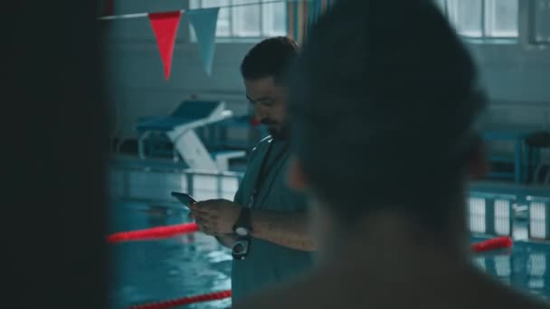 若い男性の水泳選手が部屋を変え プールに立っている中年のアジア人コーチに挨拶し スマートフォンで練習プランを見せ 指示を与える — ストック動画