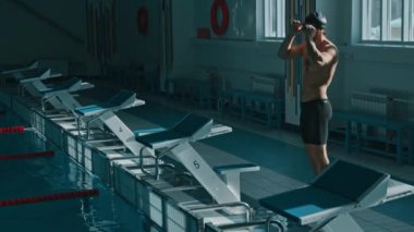Şortlu uzun boylu erkek yüzücünün tam pozu, şapka natatoriumda bilardoya gidiyor, gözlükleri ayarlıyor, dalış bloğunda duruyor, eğiliyor, suya atlamak için pozisyon alıyor.