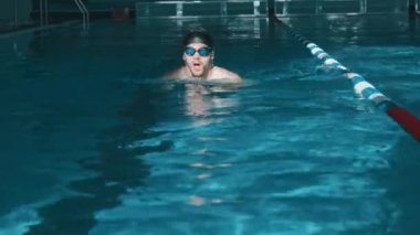 Siyah kauçuk şapkalı ve gözlüklü profesyonel sporcunun kurbağalama stilinde kameraya doğru yüzüşünü, nefesler arasında kafasının suda kayboluşunu.