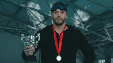 Siyah şapkalı, gözlüklü, kapüşonlu, altın madalyalı, elinde kupa, havuzda poz veren ve kameraya gülümseyen beyaz erkek yüzücünün orta boy yakın çekimi.