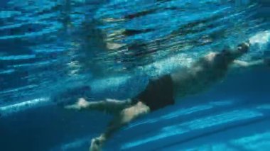 Tam eğim açısı su altında yavaş. Siyah şortlu, lastik şapkalı ve gözlüklü tanınmayan sporcunun sığ havuzun sonundaki duvara doğru serbest yüzüşü.