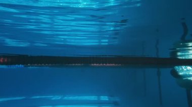 Siyah şortlu, kep ve gözlüklü, duvardan itilmiş, hava kabarcıklarından oluşan iz ile geriye doğru yüzen, su altında dönen, tanınmayan sporcunun tam yavaş sualtı çekimi.