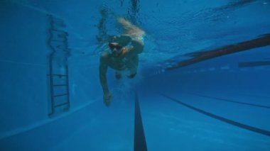 Siyah şortlu, kep ve gözlüklü, isimsiz bir adamın kucağın sonundaki havuz duvarına doğru sürünerek yaklaşırken tam sualtı görüntüsü.