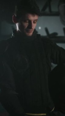Siyah üniformalı genç silahşörün dikey portresi karanlık odada durup kameraya bakarken omuz omuza yükseliyor.