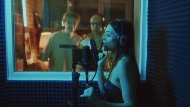 スタジオで録音をコントロールしながら 録音ブースでマイクに署名するヘッドセットのコーカサス女性ボーカリストのサイド映像 — ストック動画