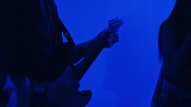 Mavi neon ışıklı stüdyoda bando eşliğinde gitar çalan ve şarkı söyleyen erkek gitaristin yakın çekimi.