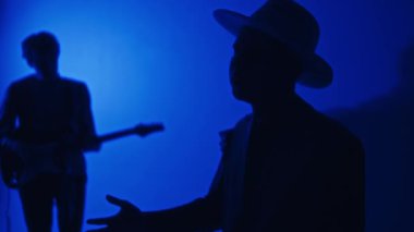 Stüdyoda neon mavisi filtreyle eşlik eden klavyeci, sentezleyici ve gitarist siluetlerinin yan yatış görüntüleri.