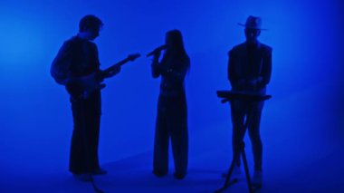 Kadın şarkıcı ve erkek gitarist ve klavyecinin mavi renkli neon filtresiyle profesyonel stüdyoda yaptıkları müzik üçlüsünün tam görüntüleri.
