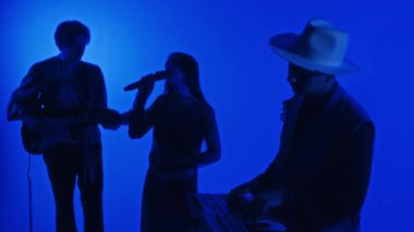 Klavyeci, gitarist ve şarkıcının belirsiz profesyonel stüdyoda mavi neon filtresiyle müzik yapmasının yan görüntüleri.