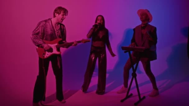 ピンクと紫色のネオンフィルターでスタジオでベースギターとシンセサイザーに伴う曲を演奏するカリスマ的なコーカサス グラム ロックバンドのフル映像 — ストック動画