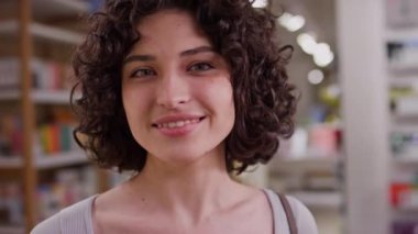 Kozmetik mağazasının önünde elinde mallarla dikilen kıvırcık kumral saçlı, mutlu bir gülümsemeyle kameraya poz veren beyaz bir kadının yakın plan portresi.