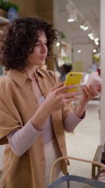 Kafkas saçlı genç bir kadının eczanede dikey görüntüsü. Elinde bir kavanoz yüz kremi var. Etiketine bakıyor, akıllı telefonda marka yazıyor.