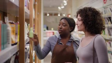 Önlüklü Afrikalı Amerikalı kadın satış asistanı beyaz bir kadınla konuşurken çekilmiş orta boy bir fotoğraf. Yeni bir cilt ürünü öneriyor. Faydasını ve kullanım yöntemini anlatıyor.