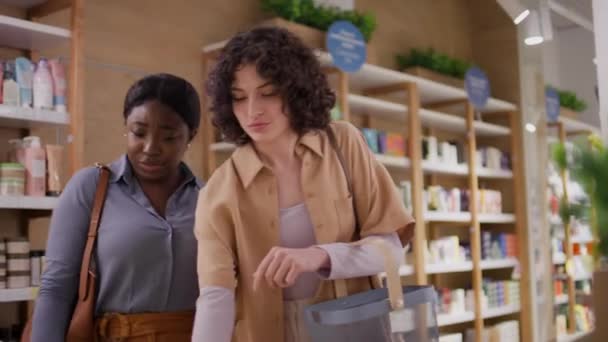 两名年轻的非洲裔美国人和白人妇女一起穿过百货商店或化妆品精品店 拿起一罐奶油 阅读标签和讨论好处的中景照片 — 图库视频影像