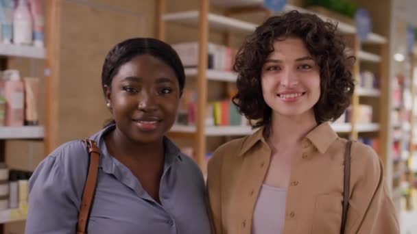 コカシアンとアフリカ系アメリカ人の2人の若い女性顧客のミディアムクローズアップポートレートショットは 商品とカメラのための笑顔で棚の前に化粧品店で一緒に立っています — ストック動画